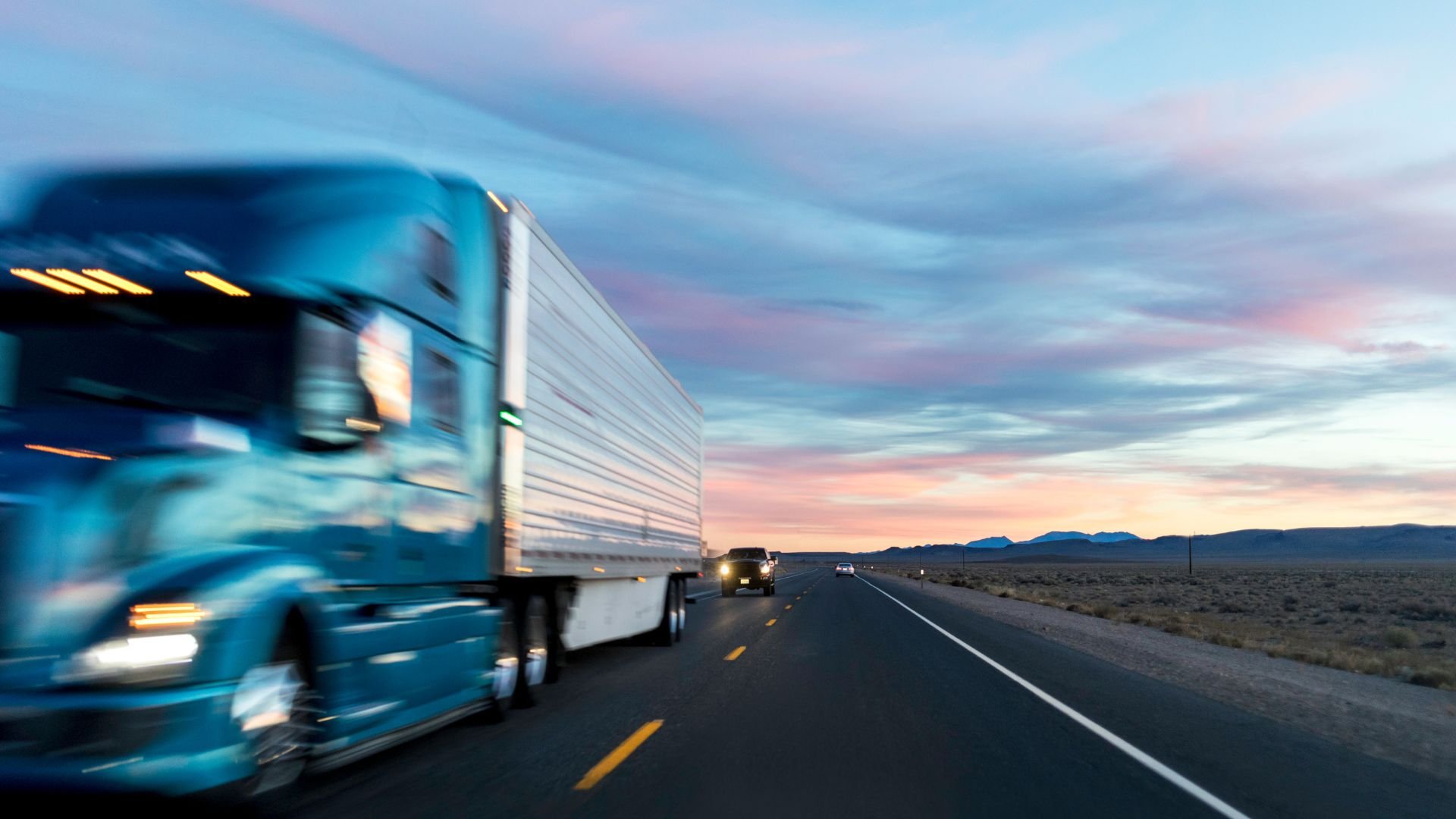 Blue semi-truck driving on highway at sunset with desert landscape